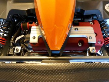 Close-up van de motorruimte van een levendig oranje en rode Ariel Atom-sportwagen, met gedetailleerde mechanische en elektronische componenten.