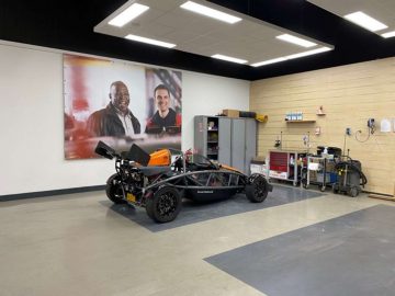 Een sportwagen, die lijkt op een Ariel Atom, in een nette werkplaats met gereedschap en een grote poster met twee lachende mannen aan de muur.