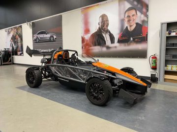 Een Ariel Atom-sportwagen in zwart en oranje, gestationeerd in de garage van een monteur, met grote portretten van twee lachende mannen aan de muur op de achtergrond.