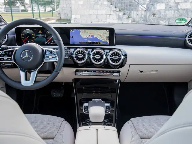 Binnenaanzicht van een Mercedes-Benz-voertuig met het stuur, het dashboard met digitale displays en crèmekleurige lederen stoelen.