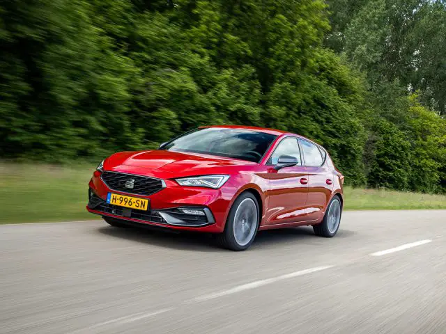 Een rode Seat Leon hatchback rijdt over een met bomen omzoomde weg.