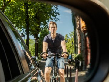 Gezicht op een jonge man op de fiets, gezien door de zijspiegel van een auto in een zonnige straat, met duidelijk de zinsnede "Zie jij mij zitten".