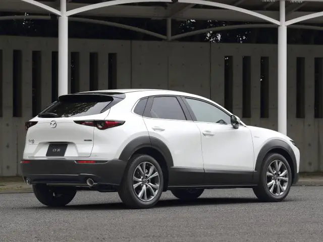 Een witte Mazda CX-30 SUV, buiten geparkeerd onder een gestructureerde overkapping, gezien vanuit de achterste driekwarthoek.