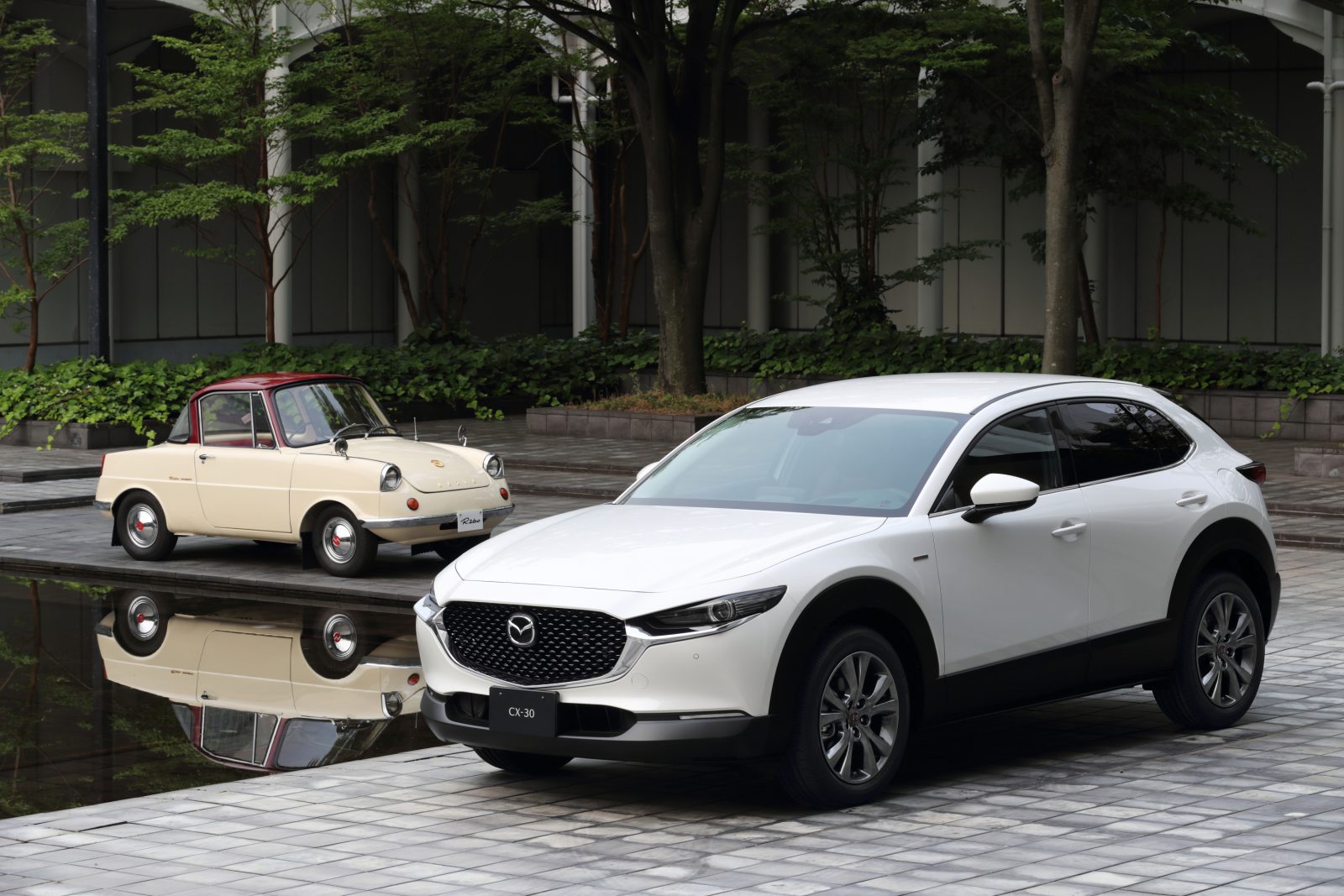 Een moderne Mazda CX-30 en een vintage crèmekleurige compacte auto geparkeerd naast een reflecterend wateroppervlak in een stadsstraat.