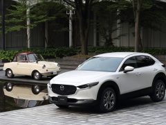 Een moderne Mazda CX-30 en een vintage crèmekleurige compacte auto geparkeerd naast een reflecterend wateroppervlak in een stadsstraat.
