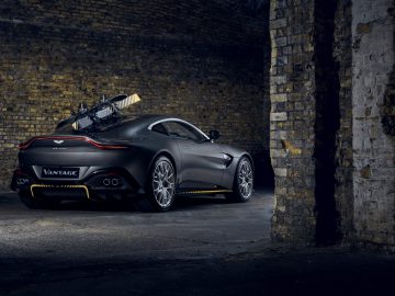 Een donkergrijze Aston Martin Vantage 007 Edition geparkeerd onder een boog, met een surfplank op het dak gemonteerd.