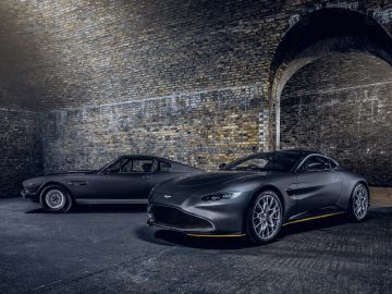 Twee luxe sportwagens, een vintage en een moderne Aston Martin 007 Edition, staan 's nachts naast elkaar geparkeerd onder een bakstenen boog.