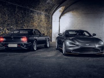 Twee auto's, een vintage model en een moderne Aston Martin 007 Edition, naast elkaar geparkeerd onder een gebogen bakstenen tunnel, verlicht door gedimd licht.