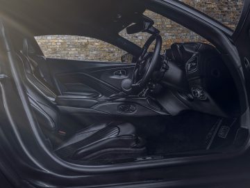 Binnenaanzicht van een Aston Martin 007 Edition met zwartleren stoelen, stuur en dashboard met bakstenen muurachtergrond door een open deur.
