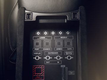 Binnenaanzicht van een Aston Martin 007 Edition met een hightech bedieningspaneel met gelabelde knoppen zoals 'laser', 'raket' en 'stargig', wat geavanceerde of fictieve functionaliteiten impliceert.