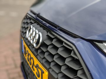 Close-up van de grille van een Audi A3-auto met het logo, met details van het zeshoekige patroon en een deel van de kentekenplaat.