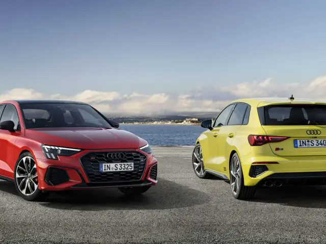 Twee Audi S3-auto's, een rode sedan en een gele hatchback, naast elkaar geparkeerd op een kustweg met een heldere hemel op de achtergrond.