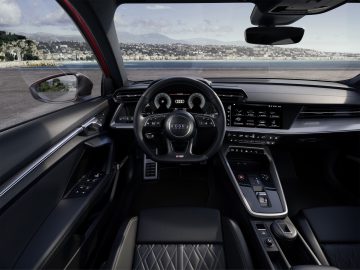 Binnenaanzicht van een Audi S3-auto vanuit het perspectief van de bestuurder, met het dashboard, het stuur en de leren stoelen, met aan de buitenkant een kustlandschap zichtbaar.