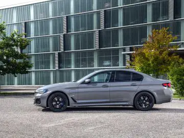 Een moderne BMW 545e xDrive sedan geparkeerd op een betonnen ondergrond met een glazen gebouw met patroon op de achtergrond.