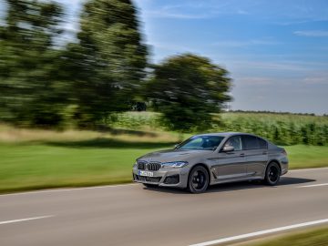 Een grijze BMW 545e xDrive snelt over een landelijke weg met wazig groen op de achtergrond, wat beweging laat zien.