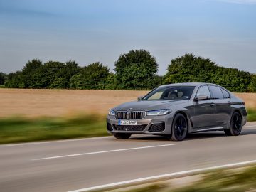 Een grijze BMW 545e xDrive sedan rijdend op een snelweg met een helderblauwe lucht en gouden velden op de achtergrond.