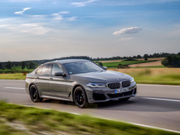 Een BMW 545e xDrive luxe sedan rijdt snel over een landelijke weg, met groene velden en een blauwe lucht op de achtergrond.