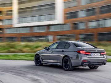 Een grijze BMW 545e xDrive sedan rijdt met hoge snelheid langs een modern gebouw.