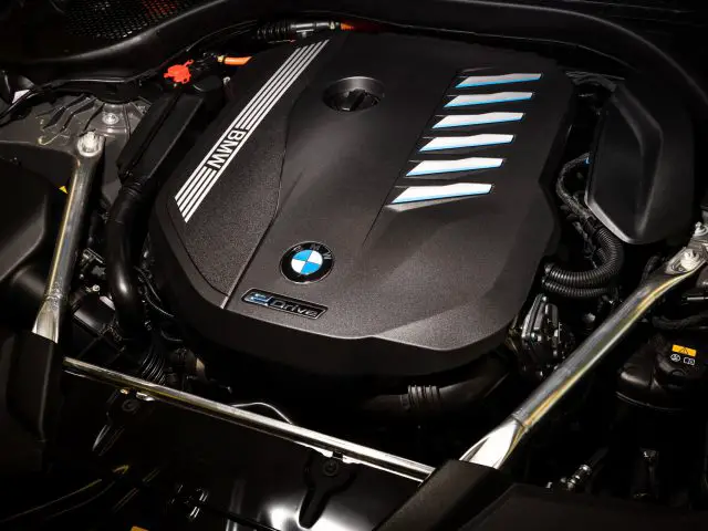 Een close-up van de motorruimte van een BMW 545e xDrive, met een strakke zwarte motorkap met het BMW-logo en blauwe accenten.