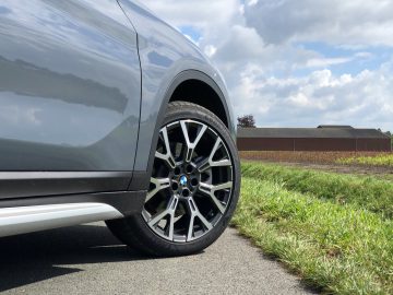 Close-up van het achterwiel van een BMW X1 xDrive25i met blauw-wit logo, geparkeerd langs een weg met uitzicht op het platteland op de achtergrond.
