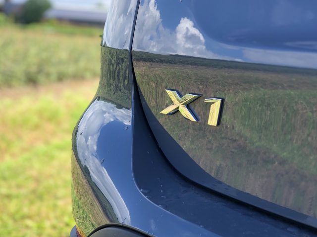 Close-up van het achterste zijpaneel van een blauwe BMW X1 met het modelbadge "xDrive25i" met een wazig groen veld op de achtergrond.