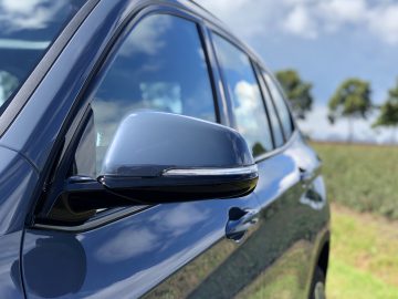 Zijaanzicht van een blauwe BMW X1 xDrive25i met focus op de spiegel, geparkeerd naast een veld onder een heldere hemel.