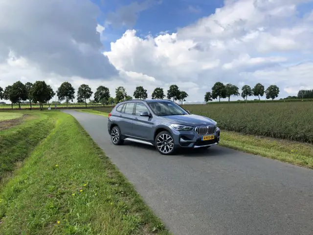 Een grijze BMW X1 xDrive25i geparkeerd op een plattelandsweg met groene velden en bomen onder een bewolkte hemel.