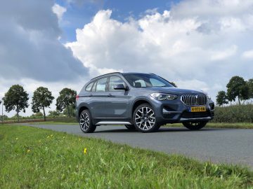 Een grijze BMW X1 xDrive25i geparkeerd op een landelijke weg met weelderige groene velden en blauwe lucht op de achtergrond.
