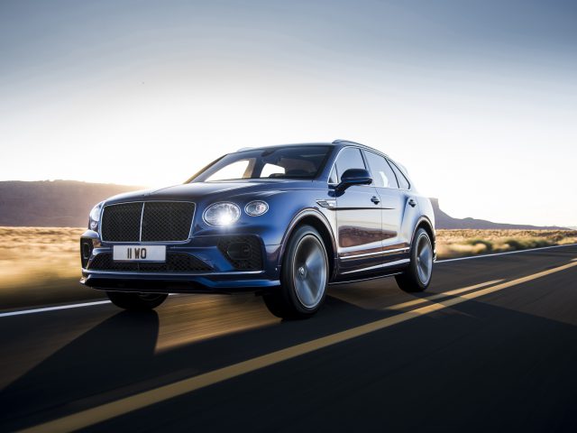 Een blauwe Bentley Bentayga Speed die op een snelweg rijdt tijdens zonsondergang, met een heldere lucht en bergen op de achtergrond.