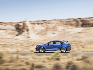 Een blauwe Bentley Bentayga Speed die over een woestijnweg rijdt met rotsachtige kliffen op de achtergrond.