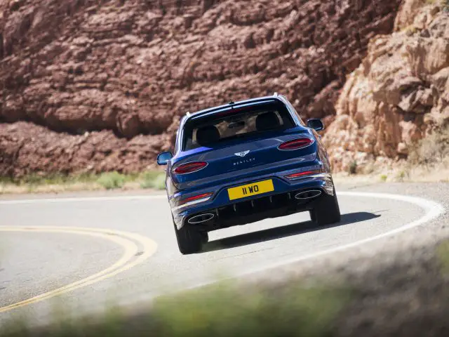 Een blauwe Bentley Bentayga Speed die over een bochtige weg rijdt met een rotsachtige heuvel op de achtergrond.