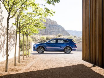 Een Bentley Bentayga Speed geparkeerd tussen houten lattenwanden met bomen en een rotsachtige heuvel op de achtergrond.