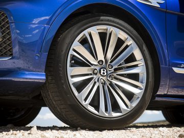 Close-up van een blauwe Bentley Bentayga Speed-lichtmetalen velg met het Bentley-logo op de wieldop, geparkeerd op een grindoppervlak.