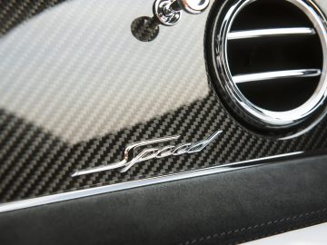 Close-up van een 'Bentley Bentayga Speed'-embleem op een koolstofvezelpaneel met daarboven een chromen ronde ventilatieopening.