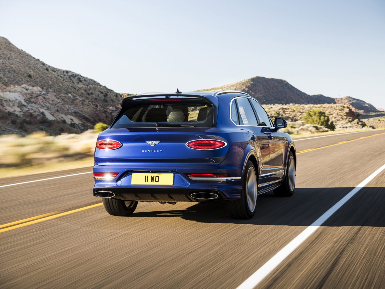 Een blauwe Bentley Bentayga Speed SUV die op een snelweg door een woestijnlandschap rijdt.