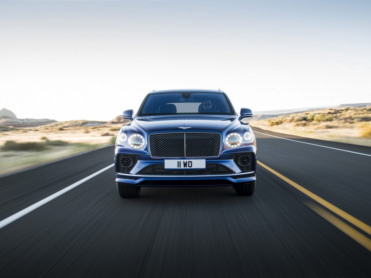 Een blauwe Bentley Bentayga Speed die te hard rijdt op een open snelweg met een heldere lucht en een woestijnlandschap op de achtergrond.