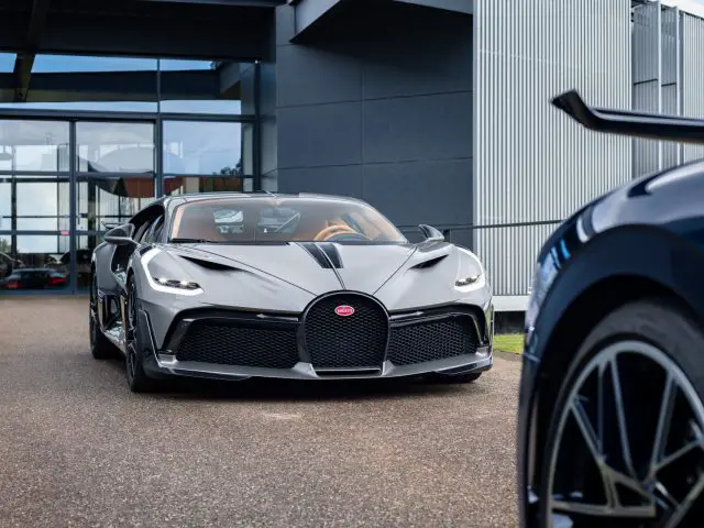 Een Bugatti Divo geparkeerd voor een modern gebouw, met aan de rechterkant gedeeltelijk een andere luxe auto zichtbaar.