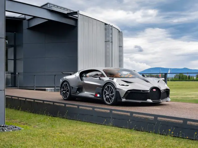 Een zilveren Bugatti Divo geparkeerd buiten een modern gebouw met een bewolkte blauwe lucht op de achtergrond.