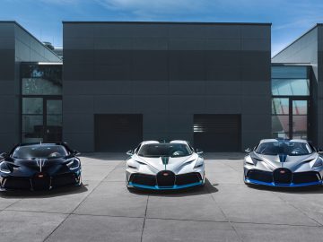 Drie Bugatti Divo-sportwagens, in het zwart, groenblauw en zilver, geparkeerd voor een modern, donker gebouw onder heldere hemel.