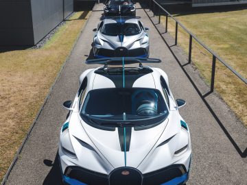 Luchtfoto van vijf luxe sportwagens, waaronder een Bugatti Divo, in afwisselend witte en blauwe kleuren, geparkeerd in een rij tussen moderne gebouwen.