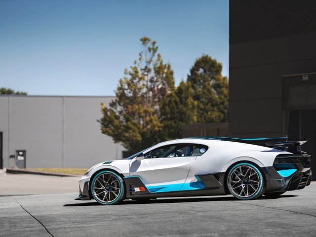 Een wit-blauwe Bugatti Divo parkeerde op een open plek naast een gebouw, onder een heldere hemel.