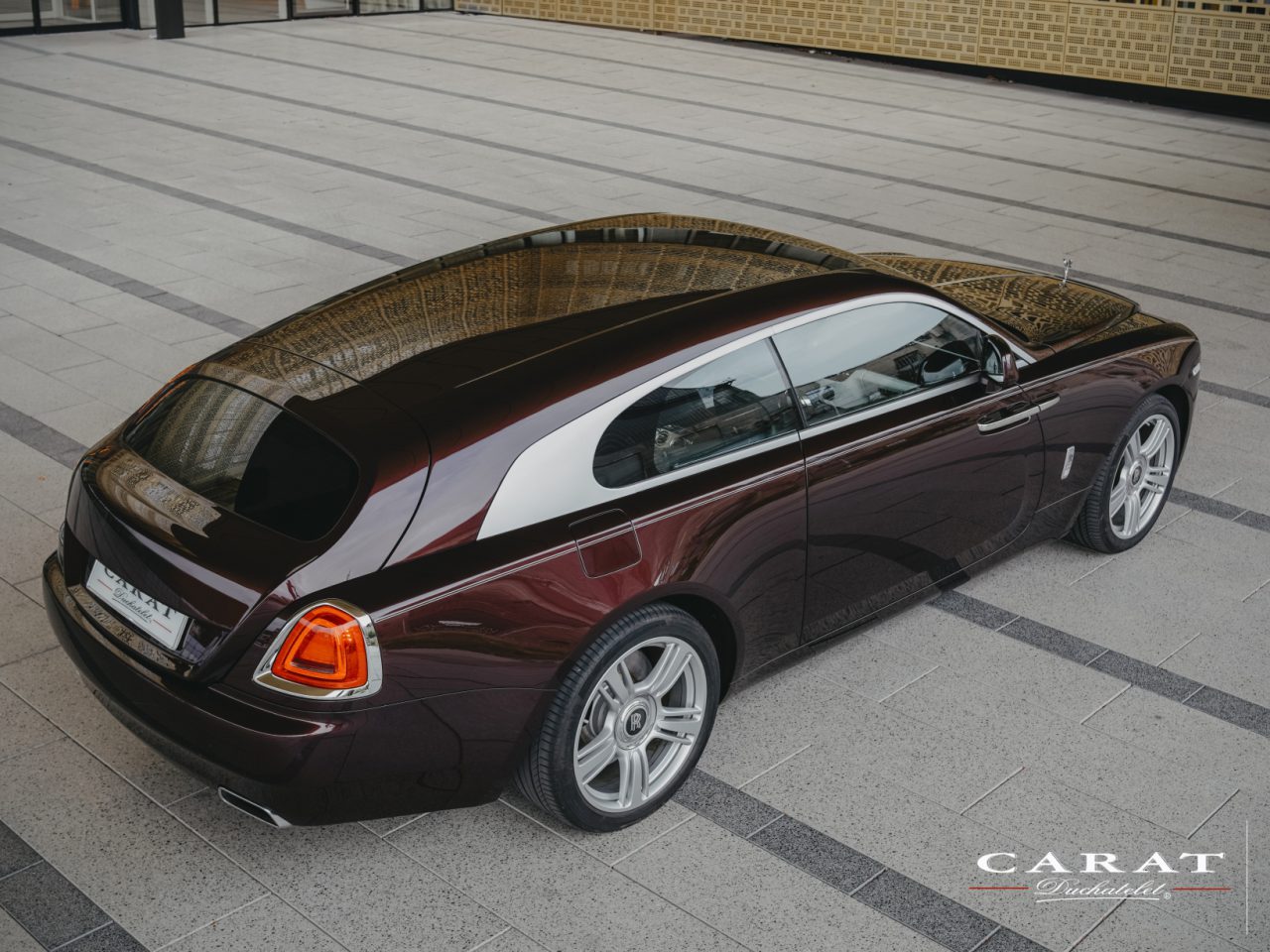 Luchtfoto van een kastanjebruine Rolls-Royce Silver Spectre Shooting Brake met een uniek golvend goud en zwart dak geparkeerd op een binnenplaats.