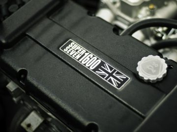 Close-up van een zwarte motorkap met een "Caterham Super Seven 1600" sticker en een Union Jack-embleem, tegen een onscherpe motorachtergrond.