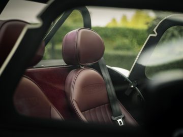 Het interieur van een luxe Caterham Super Seven 1600-auto, met de nadruk op de karmozijnrode lederen stoelen met geborduurde logo's, gezien door een open deur.