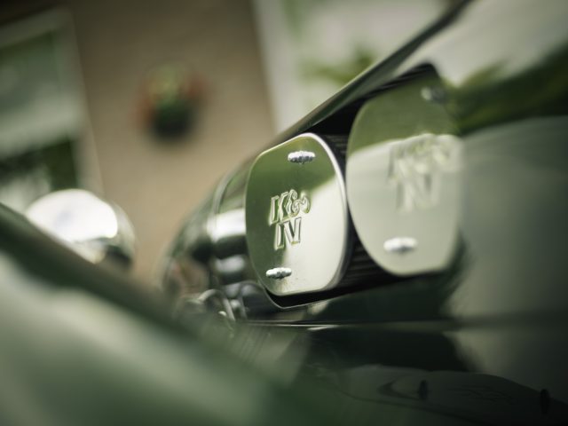 Close-up van twee groene Caterham Super Seven 1600 motorkapemblemen met zilverchromen details, die een wazige groene en witte achtergrond weerspiegelen.