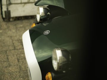 Close-up van de voorkant van een groen-witte Caterham Super Seven 1600, met ronde koplampen en een glanzend embleem.