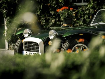 Vintage groene Caterham Super Seven 1600 buiten geparkeerd, gedeeltelijk verborgen door gebladerte op de voorgrond, met de nadruk op de voorkant en de grille.