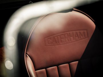 Een close-up van een bruinleren Caterham Super Seven 1600 autostoeltje met de merknaam in reliëf.