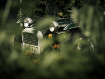 Een vintage groene Caterham Super Seven 1600 sportwagen, gedeeltelijk verborgen door wazig gebladerte op de voorgrond.