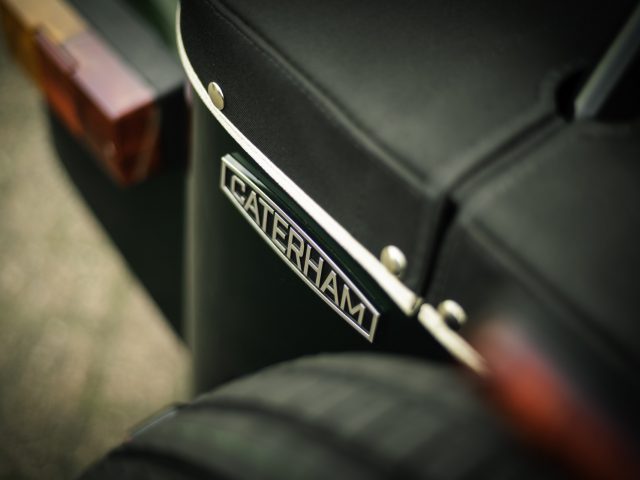 Close-up van het embleem van een Caterham Super Seven 1600-auto op de zijkant van een zwartleren stoel, met een onscherpe achtergrond.